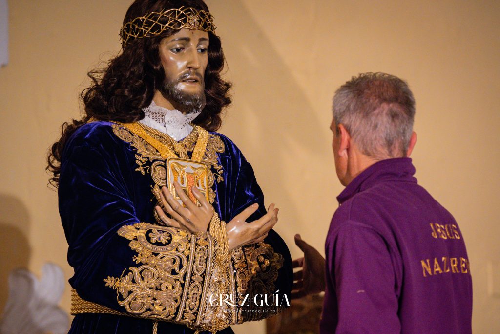 Primer plano de la imagen de Nuestro Padre Jesús Nazareno, con detalles de su vestimenta, mientras el vestidor permanece en segundo plano. Fotografía de La Cruz de Guía – Ikonox.
