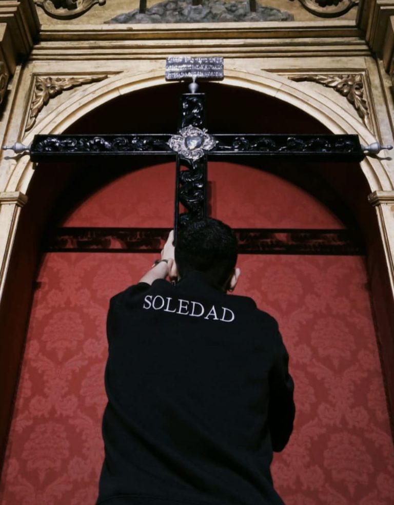 Joven hermano de la Cofradía de Nuestra Señora de la Soledad de Alcázar de San Juan sosteniendo una cruz, con camiseta que lleva el nombre "Soledad", fotografía de Ikonox.